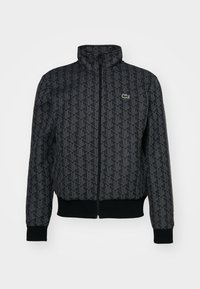 Veste noire zippée avec motif géométrique sur toute la surface, poignets et ourlet côtelés, petit logo crocodile vert sur le côté gauche de la poitrine.