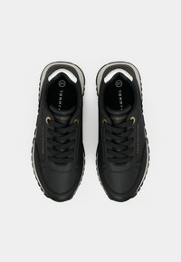 Svarte sneakers med en glatt skinnoverdel, rund tå og kontrasterende grå såle. Gullakenter på snøringen; logo på tungen og siden.