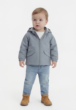 Maluch ubrany w szarą kurtkę z kapturem, białą koszulkę, jasnoniebieskie dżinsy i beżowe buty na sznurowadła, stojący na białym tle.