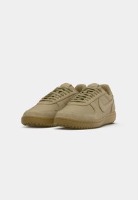 Een paar beige lage sneakers met veters, een gestructureerde zool en gestikte details, geplaatst op een wit oppervlak.