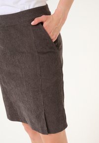 Kvindes hånd i lommen på en mørkegrå, knælang fløjlsslag med sideslids, iført en hvid top.