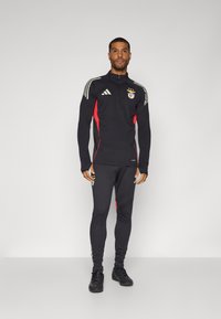 Musta urheiluasu punaisilla yksityiskohdilla, jossa on vetoketjullinen pusero ja tiukat housut. Sisältää Adidas-logon ja joukkueen embleemin.
