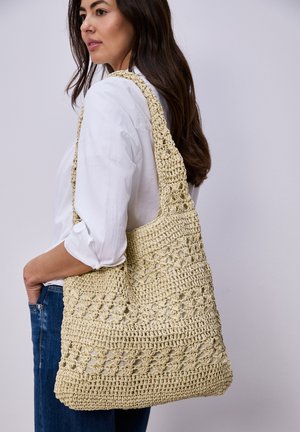 Donna con capelli scuri che indossa una camicia bianca e jeans porta una grande borsa beige all'uncinetto a tracolla su sfondo uniforme.