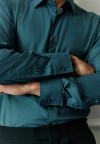 Teal long-sleeve shirt with a smooth texture, vertical stripes, and buttoned cuffs, worn with a black waistband.