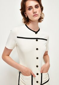 Weißes kurzes Kleid mit kurzen Ärmeln und schwarzen Akzenten, das große schwarze Knöpfe und Seitentaschen aufweist, mit glatter Textur und figurschmeichelndem Design.
