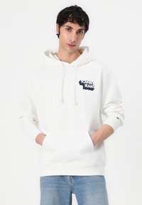 Hoodie blanc en mélange de coton doux avec une poche kangourou à l'avant. Présente un écusson logo bleu marine sur la poitrine et des poignets côtelés.