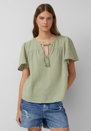 Jeune femme aux longs cheveux bruns portant un chemisier vert clair à manches courtes et un short en jean bleu déchiré sur un fond uni.
