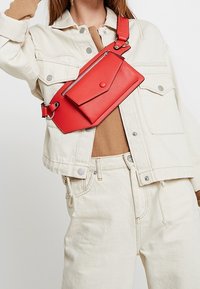 Borsa a tracolla rossa in ecopelle con tasca con zip e chiusura a scatto. Presenta una tracolla regolabile e una forma rettangolare.