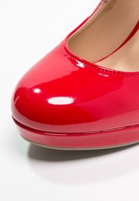 Talons hauts en cuir verni rouge, design à bout rond, finition glossy, détail de couture subtil le long du bord, semelle plate avec légère élévation.