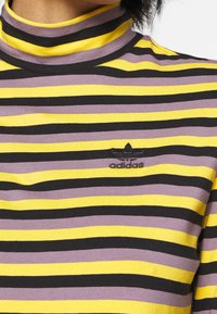 Haut à col roulé rayé avec des rayures horizontales violettes, jaunes et noires. Présente un petit logo adidas noir au niveau de la poitrine.