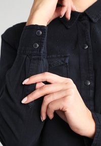 Personne ajustant le poignet d'une chemise à manches longues noire avec des boutons pression et une poche avant visible.
