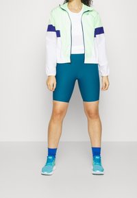 Jasnozielona i biała kurtka przeciwdeszczowa z fioletowymi paskami, biały podkoszulek, turkusowe spodenki rowerowe oraz niebieskie buty sportowe z pasującymi skarpetkami.