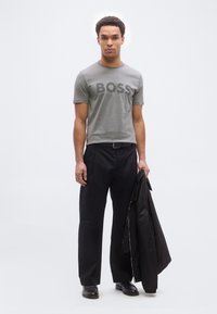 Άνδρας που φοράει γκρι μπλουζάκι BOSS, μαύρο παντελόνι με φαρδιά πόδια, μαύρα παπούτσια, κρατώντας μαύρο σακάκι μπροστά σε λευκό φόντο.