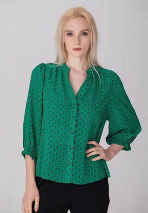 Femme blonde aux cheveux raides porte un chemisier vert à motifs avec des manches bouffantes et un pantalon noir, posant avec une main sur la hanche.
