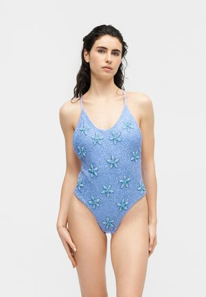 Femme portant un maillot de bain une pièce bleu clair avec de fines bretelles et des motifs d'étoiles de mer, debout devant un fond blanc uni.