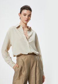 Camicia beige con bottoni in tessuto texture, maniche lunghe e colletto, abbinata a pantaloni in satin marrone chiaro a vita alta con tasche laterali.