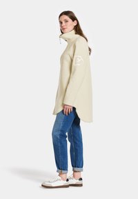 Creme fleece pullover med høj krave og slids i siderne. Har en struktureret stof og en geometrisk lap på venstre arm. Stylet med blå jeans og sko.