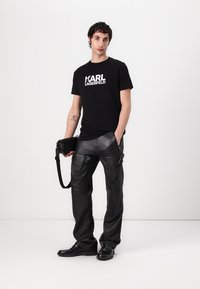 T-shirt en coton noir avec le logo blanc "KARL LAGERFELD", associé à un pantalon en cuir noir. Le modèle tient un sac bandoulière noir.
