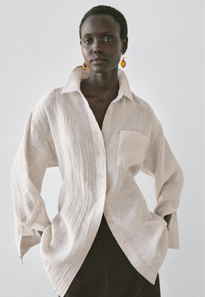 Kobieta w luźnej, teksturowanej, kremowobiałej koszuli z kołnierzykiem i guzikami, z bursztynowymi kolczykami wiszącymi, ubrana w ciemną spódnicę, stoi na tle jednolitego szarego tła.