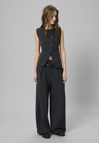 Ujjatlan, szabott sötétszürke mellény V-alakú elejével és öt gombjával, amelyet magas derekú, bő szárú sötét nadrággal és fekete cipővel viselnek.