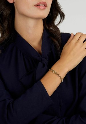 Bracciale d'oro con anelli circolari e accentuazioni decorative indossato su un polso nudo, abbinato a una blusa blu navy con scollo legato.
