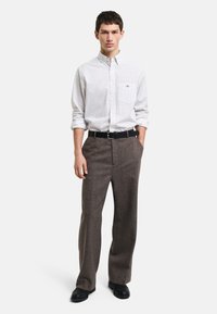 Camisa blanca de cuello con pequeños lunares azules, pantalones marrones texturizados, cinturón de cuero negro y zapatos negros. El modelo está de pie con las manos en los bolsillos.