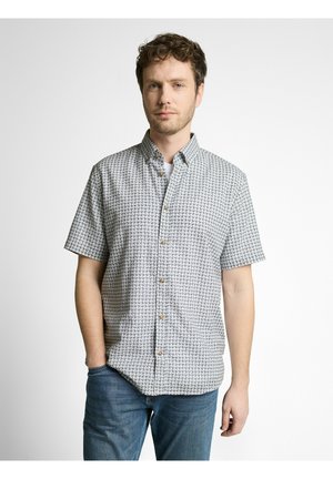 Mann mit kurzen lockigen Haaren, der ein gemustertes kurzärmeliges Hemd und blaue Jeans trägt, steht mit einer Hand in der Tasche.