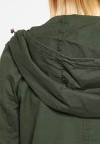 Chaqueta verde oliva con capucha, costuras visibles y detalles metálicos en el cuello. La tela parece suave con una textura mate.