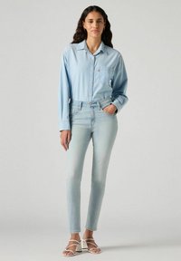 Camisa de mezclilla azul claro con cierre de botones y bolsillo, combinada con jeans ajustados de lavado claro. Sandalias blancas con tiras completan el conjunto.