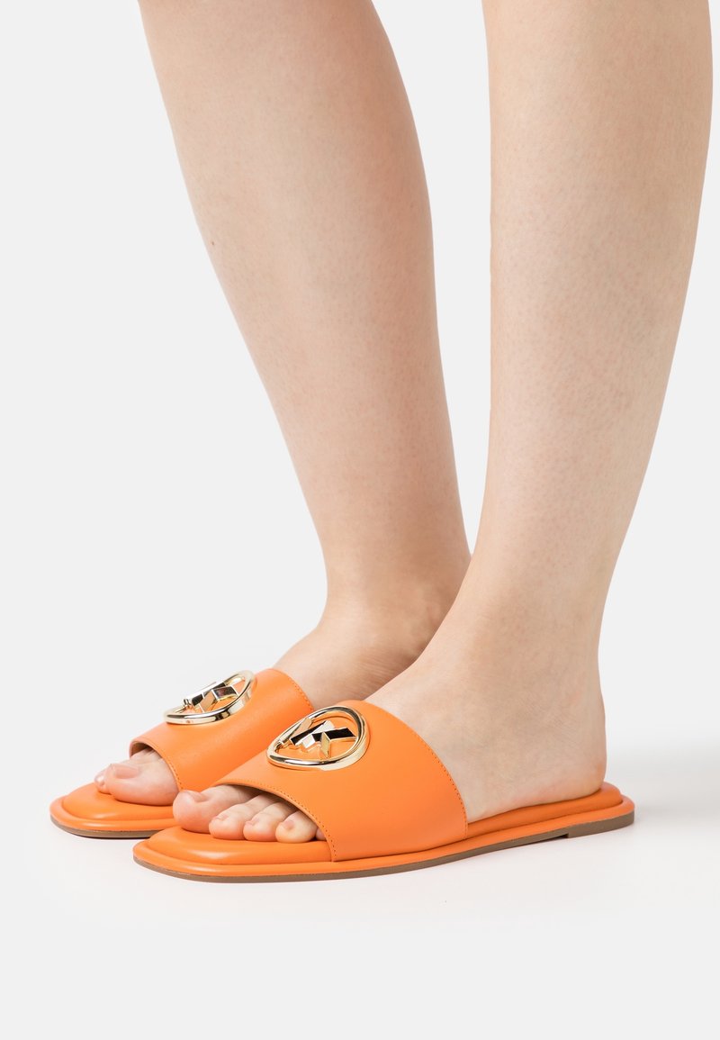Pieds portant des sandales slides orange avec des embellissements circulaires dorés sur le dessus, sur un fond clair uni.