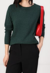 Femme portant un pull en maille vert foncé et un pantalon noir avec un sac à bandoulière rouge, main reposant dans la poche, devant un fond uni.