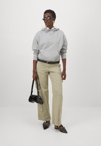Felpa grigia con tasca e coulisse, pantaloni cargo beige a gamba larga con zip, borsa nera in vernice e décolleté nere a punta.