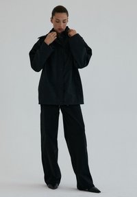 Kobieta poprawiająca kołnierz czarnej, oversizedowej kurtki, ubrana w pasujące czarne spodnie o szerokich nogawkach i czarne czółenka, stojąc na jednolitym tle.