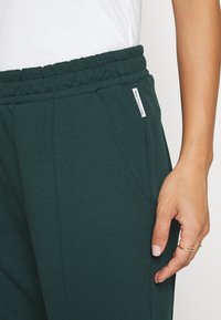 Dunkelgrüne Sweatpants aus einem weichen Baumwoll-Mix, mit einem elastischen Bund, Seitentaschen und einem kleinen Markenschild an der Seite.