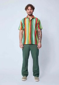 Chemise multicolore à manches courtes avec des rayures verticales en rouge, orange, vert et bleu, associée à un pantalon vert et des chaussures de couleur claire.
