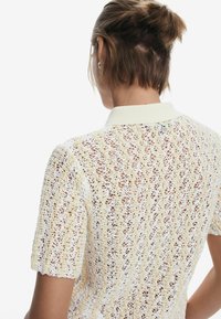 Pletená polo košeľa v krémovej farbe s čipkovaným vzorom, zdobená krátkymi rukávmi a rebrovaným golierom. Textúrovaná, ľahká tkanina.