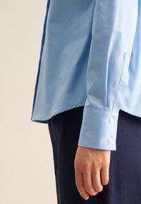 Camicia button-up azzurra chiara con maniche lunghe e polsini a un bottone, caratterizzata da un tessuto liscio e un orlo leggermente curvo.