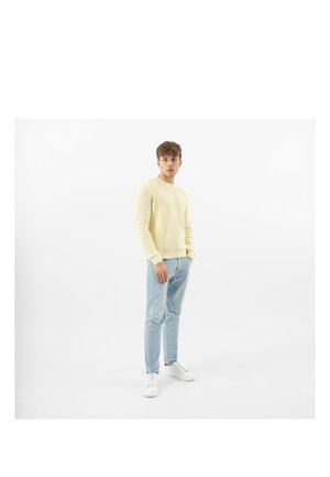 Un jeune homme se tient les mains dans les poches, portant un pull jaune pâle, un jean bleu clair et des baskets blanches sur un fond blanc.