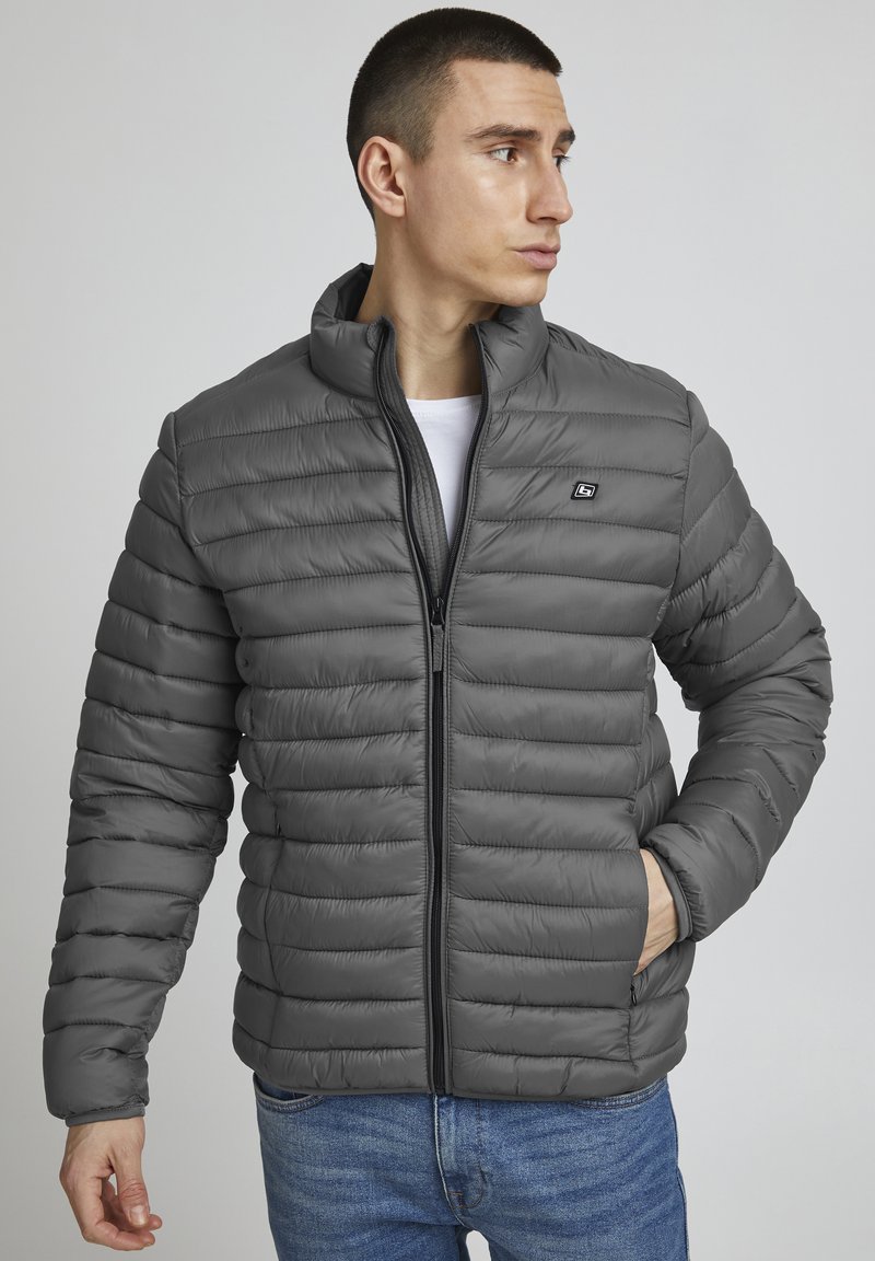 Mees, kes kannab halli tikk-kangast jopet ees oleva tõmbluku ja taskuga, vaatab kõrvale, seisab ühevärvilise hele tausta ees.