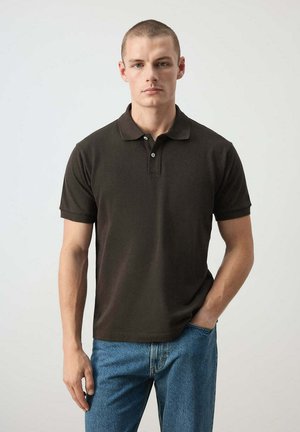 Uomo con capelli corti che indossa una polo nera e jeans blu, in piedi con una mano in tasca davanti a uno sfondo semplice.