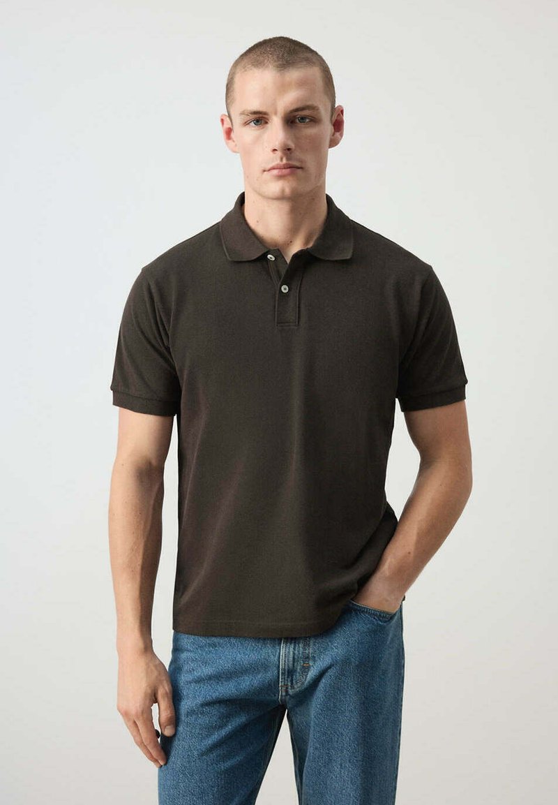Homme aux cheveux courts portant un polo noir et un jean bleu, debout avec une main dans la poche face à un fond uni.