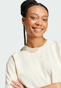 Krémová mikina s kulatým výstřihem a krátkými rukávy, zdobená jemným, texturovaným výšivkovým logem na hrudi.