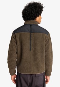 Brown fleece jacket with a black nylon panel on the shoulders, high collar, and a vertical zipper detail at the back. Textured surface.