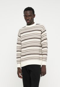 Pull en tricot crème clair avec des rayures horizontales brunes, des poignets et un ourlet côtelés, et des motifs texturés. Assorti avec un pantalon noir.