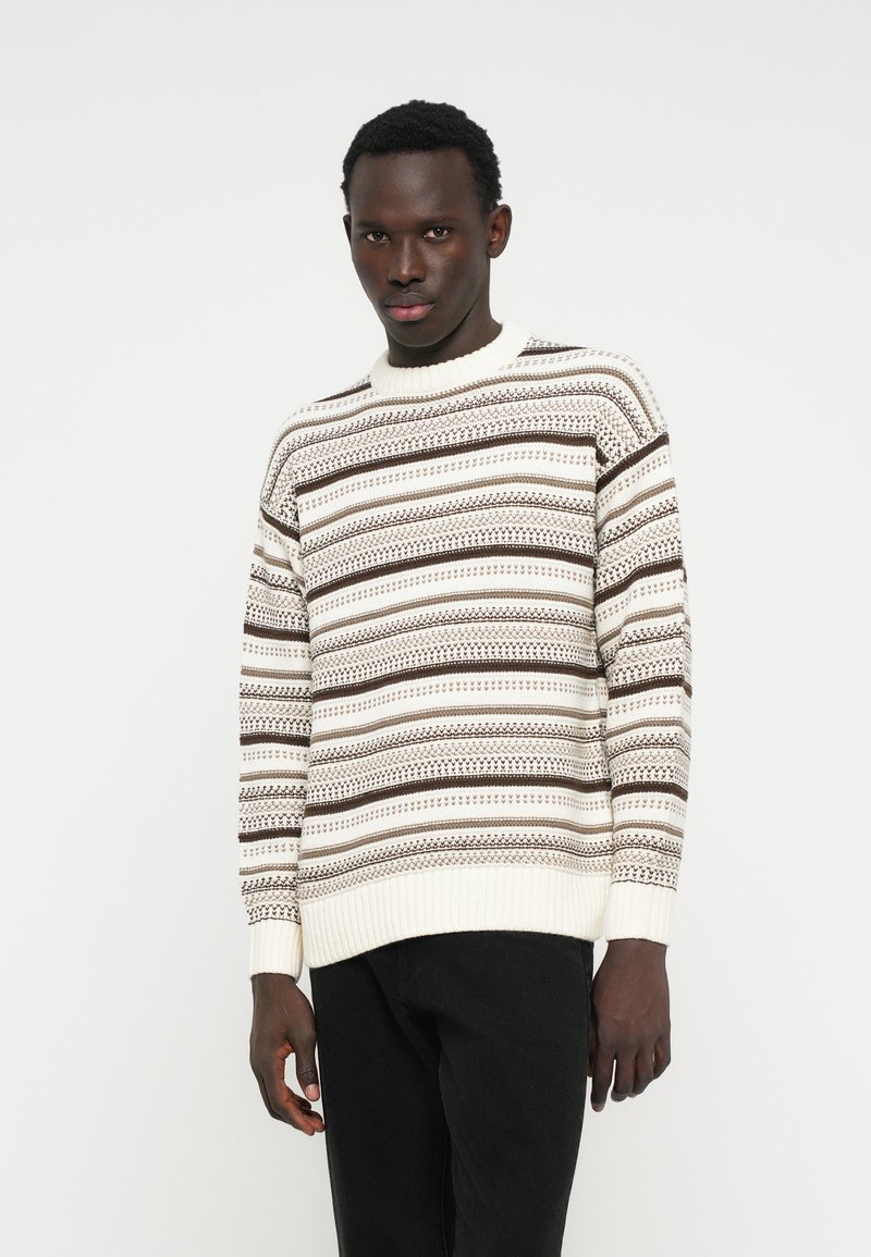 Pull en tricot crème clair avec des rayures horizontales brunes, des poignets et un ourlet côtelés, et des motifs texturés. Assorti avec un pantalon noir.