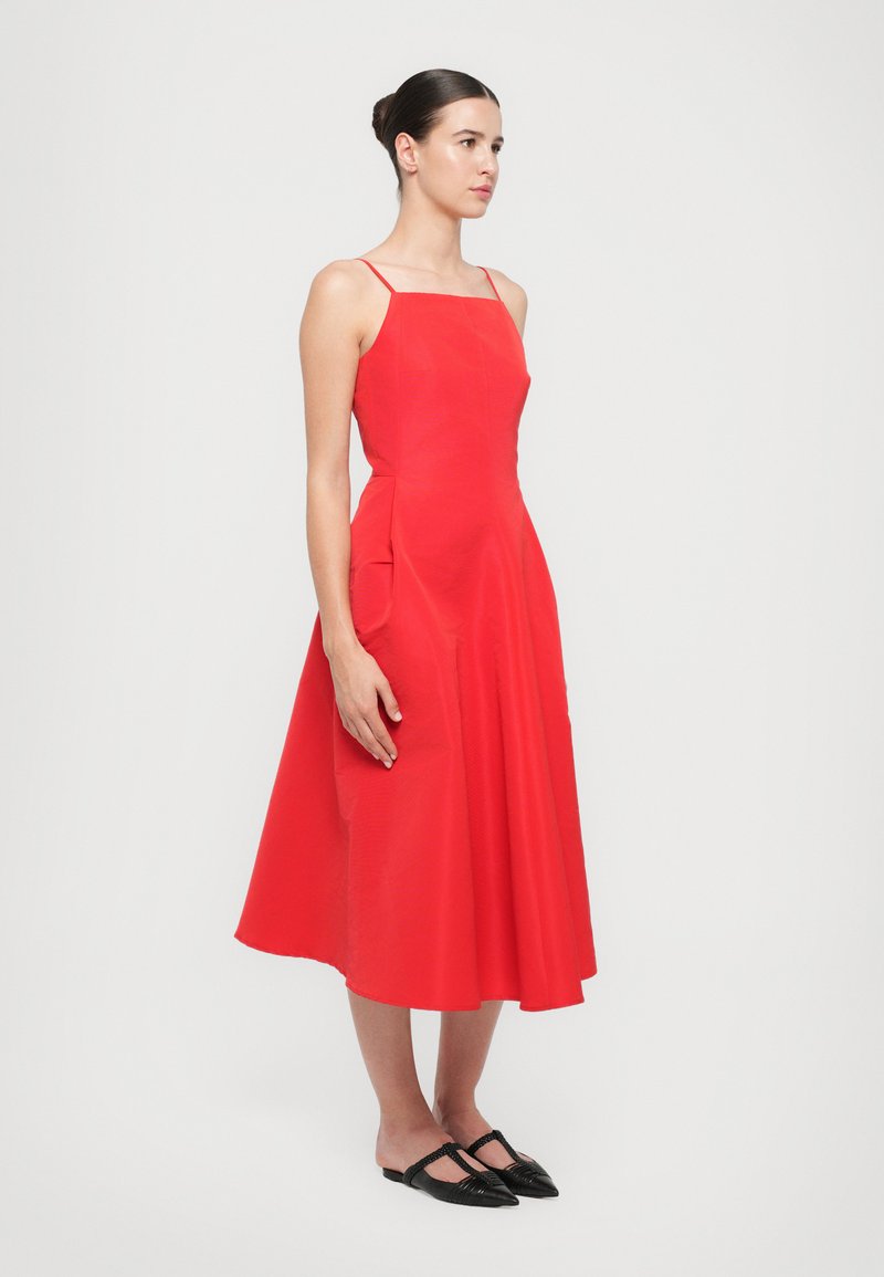 Le mannequin pose dans une robe rouge sans manches, arrivant au genou, avec des poches latérales et des chaussures plates noires, sur un fond blanc uni.