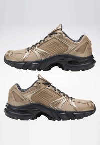 Béžové sportovní boty s texturovaným síťovým svrškem, černou podrážkou a kontrastními tmavými akcenty. Disponují šněrovacím systémem a vyztuženou konstrukcí.