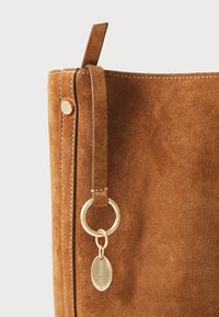 Braune Wildlederhandtasche mit glatter Textur, ausgestattet mit einem goldenen Metallring und einem Etikett sowie einem genähten Ledertragegurt.