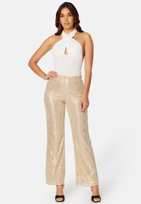 Bílý halter bodysuit má klíčovou dírku; zlaté široké kalhoty s flitry se třpytí s vertikálními vzory; model má na sobě černé sandály na podpatku.