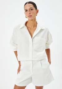 Camicia bianca in lino con bottoni, dalla vestibilità rilassata e taschino anteriore singolo, abbinata a pantaloni corti bianchi su misura, che mostrano una superficie liscia.